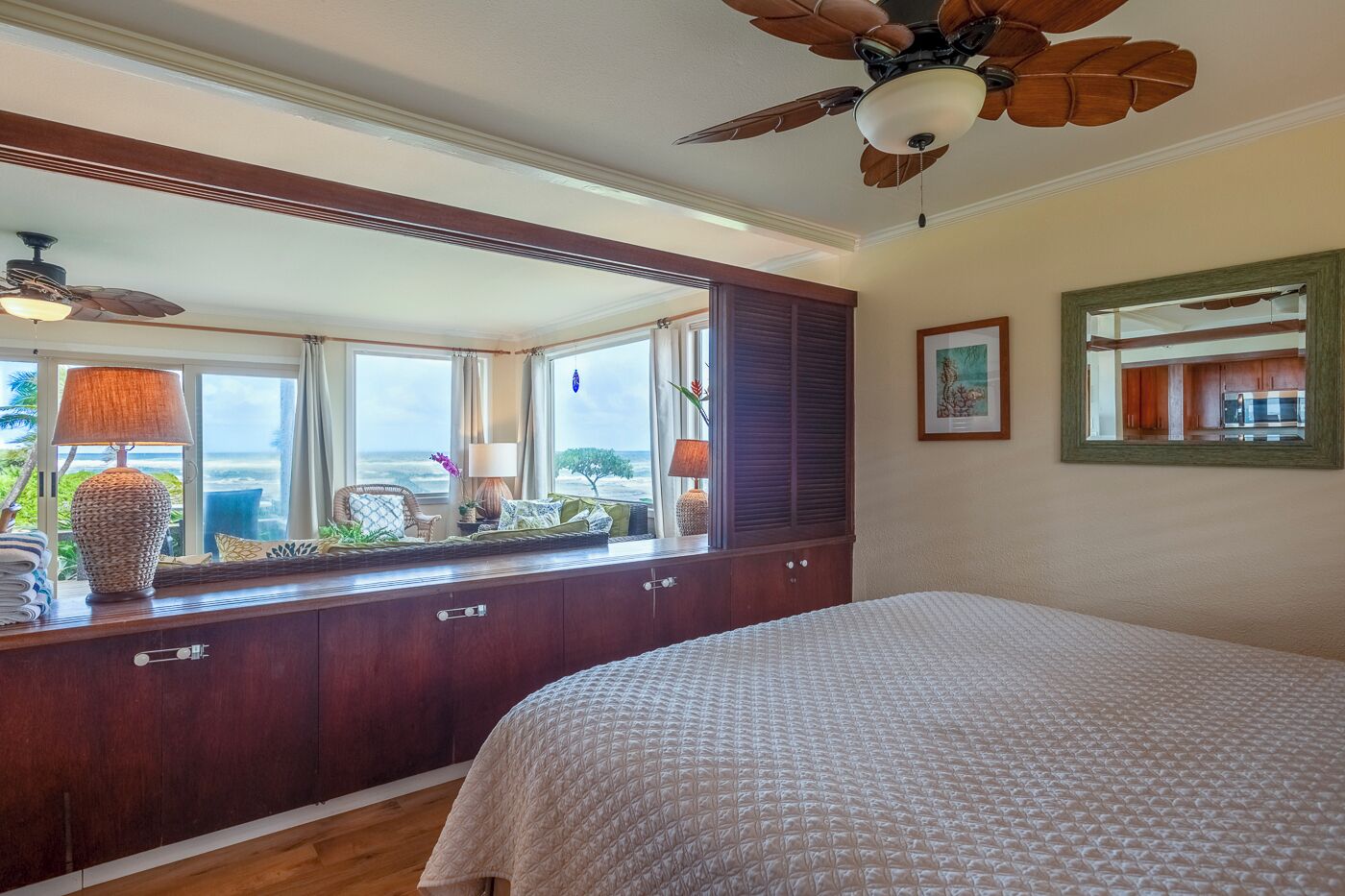 A bedroom with a large bed, a wooden dresser, a ceiling fan, and a view of the ocean through large windows.