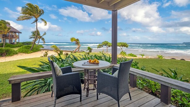 A covered outdoor seating area with a table and chairs, overlooking a beach with palm trees and ocean waves. - Hanalei Colony Resort I-1 (Hanalei)