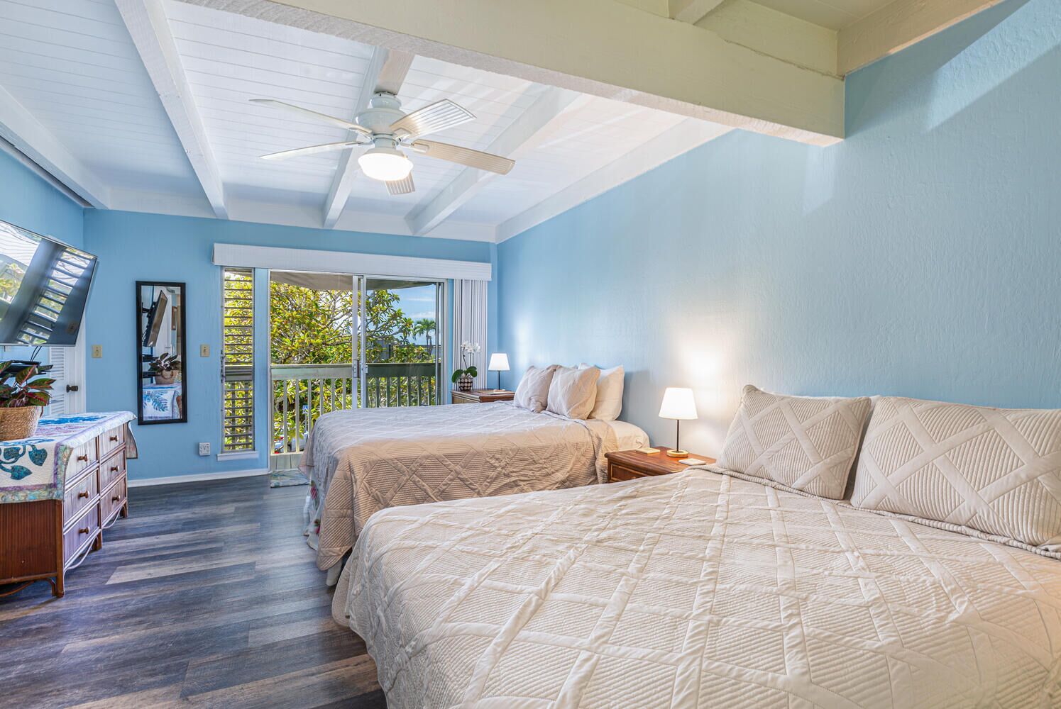 A bedroom with two beds, a ceiling fan, a large window with a view of trees, and a dresser with drawers.