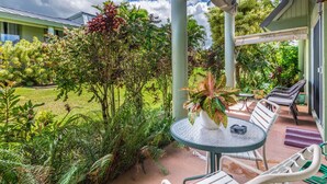 A covered outdoor seating area with a table and chairs, surrounded by lush greenery and a view of a building in the background.