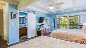 A bedroom with two beds, a ceiling fan, a TV, a bathroom with a sink and mirror, and a balcony with a view of trees.