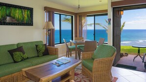 A room with a green sofa, wicker chairs, a wooden table, and a view of the ocean. - Sealodge G4 (Princeville)