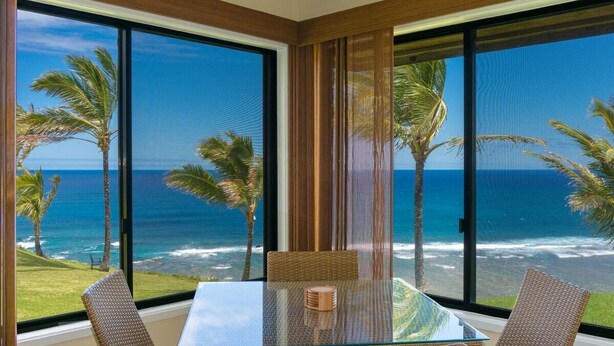 A room with a glass table and wicker chairs, overlooking a beach with palm trees and the ocean. - Sealodge G4 (Princeville)