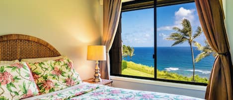 A bed with floral bedding, a lamp, and a view of the ocean through a window.