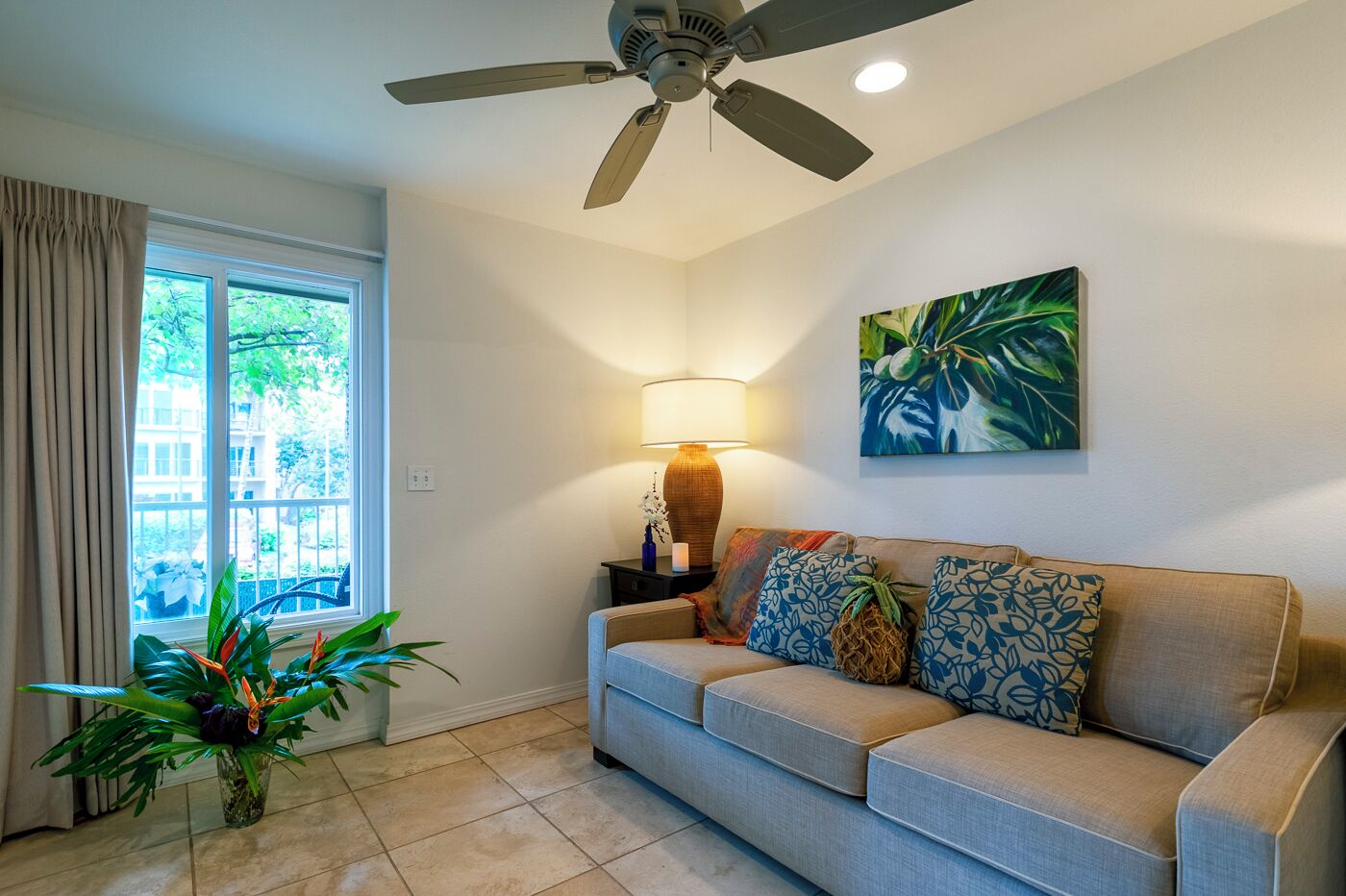 A living room with a ceiling fan, a sofa with patterned cushions, a floor lamp, a potted plant, and a painting on the wall.