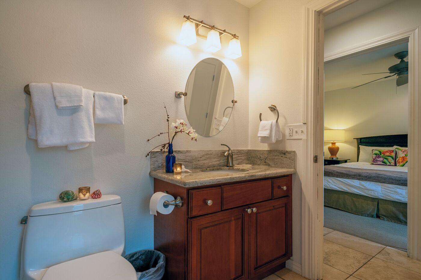 Ein Badezimmer mit Toilette, Waschbecken und Spiegel. Handtücher hängen an der Wand und ein Mülleimer steht bereit.
