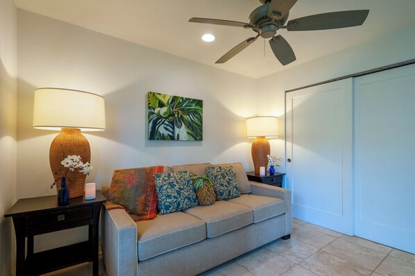 A living room with a sofa, two side tables with lamps, a ceiling fan, a door, and a wall-mounted painting. - Kauai Kailani 218 (Kapaa)