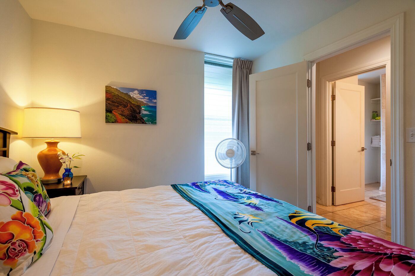 Una habitación de hotel con una cama, una mesita de noche con lámpara, un ventilador de pared, un cuadro de un paisaje costero y una puerta que da al baño.