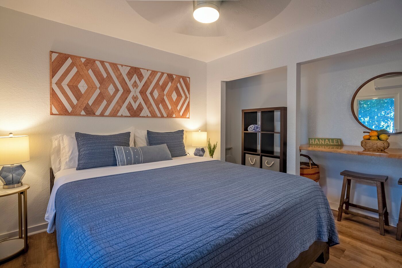 Un dormitorio con una cama grande, una mesita de noche con lámpara, un armario de madera, un espejo y un taburete pequeño.