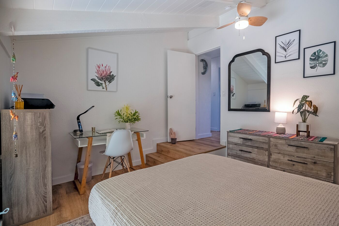 A bedroom with a bed, a desk with a chair, a dresser, a mirror, and artwork on the walls.