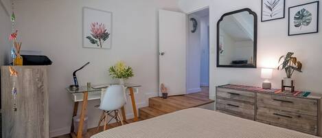 A bedroom with a bed, a desk with a chair, a dresser, a mirror, and artwork on the walls.