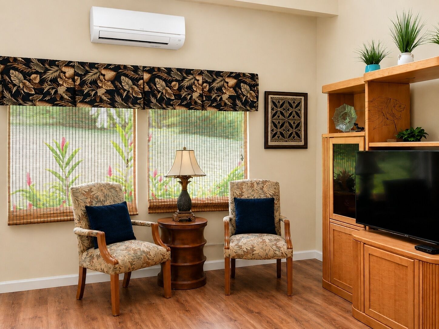 A living room with two patterned armchairs, a small table with a lamp, a television on a wooden stand, and a large window with a view of greenery.