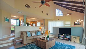 A spacious living room with a beige sofa, a wooden coffee table, and a TV stand. There are ceiling fans, a staircase, and a kitchen area visible. - Kamahana 23 (Princeville)