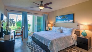 A bedroom with a bed, a ceiling fan, a painting, a nightstand, a lamp, a chair, a table, and a view of the outdoors. - Kamahana 23 (Princeville)