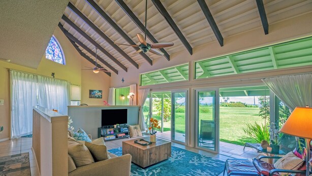 A spacious living room with a television, comfortable seating, a coffee table, and a view of a lush garden through large sliding glass doors. - Kamahana 23 (Princeville)