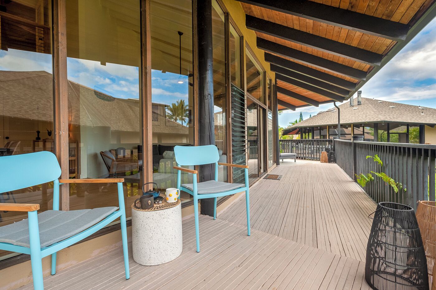 Un espacio al aire libre cubierto con piso de madera, dos sillas azul claro, una mesa redonda con tetera y taza, y vista a un edificio con techo inclinado.
