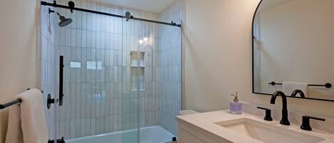 A modern bathroom with a glass shower enclosure, a white sink with black faucets, and a large mirror.