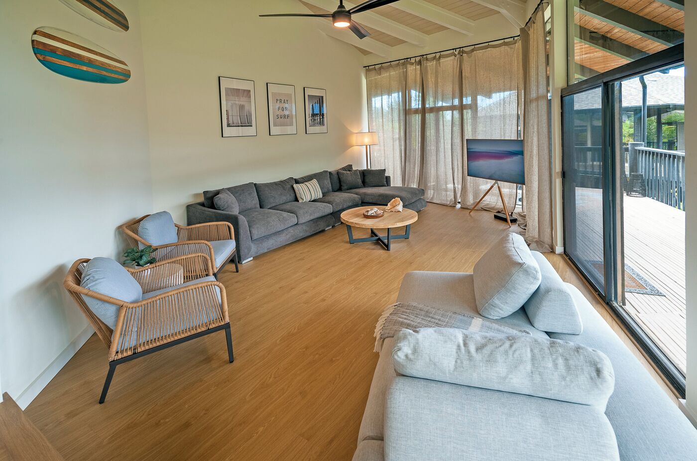 A spacious living room with a grey sofa, a wicker chair, and a round coffee table. There are three framed pictures on the wall and a large window with curtains.
