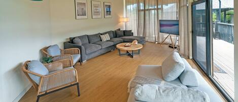A spacious living room with a grey sofa, a wicker chair, and a round coffee table. There are three framed pictures on the wall and a large window with curtains.