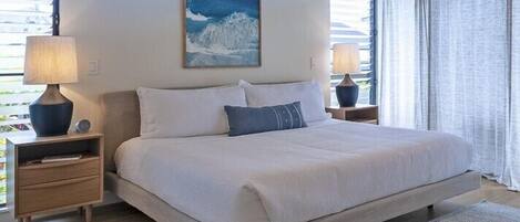 A bedroom with a large bed, two bedside tables, a nightstand, a window with blinds, and a painting on the wall.