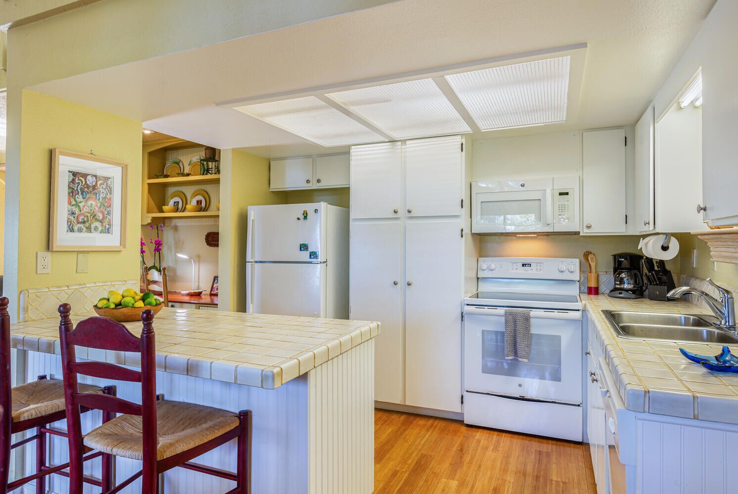 A compact kitchen with white cabinets, a refrigerator, an oven, and a microwave. There is a dining area with a chair and a table, and a fruit bowl on the counter.