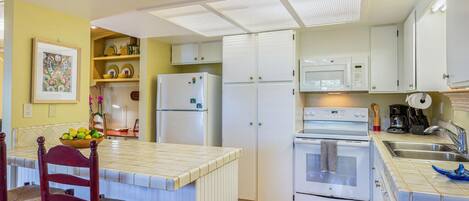 A compact kitchen with white cabinets, a refrigerator, an oven, and a microwave. There is a dining area with a chair and a table, and a fruit bowl on the counter.