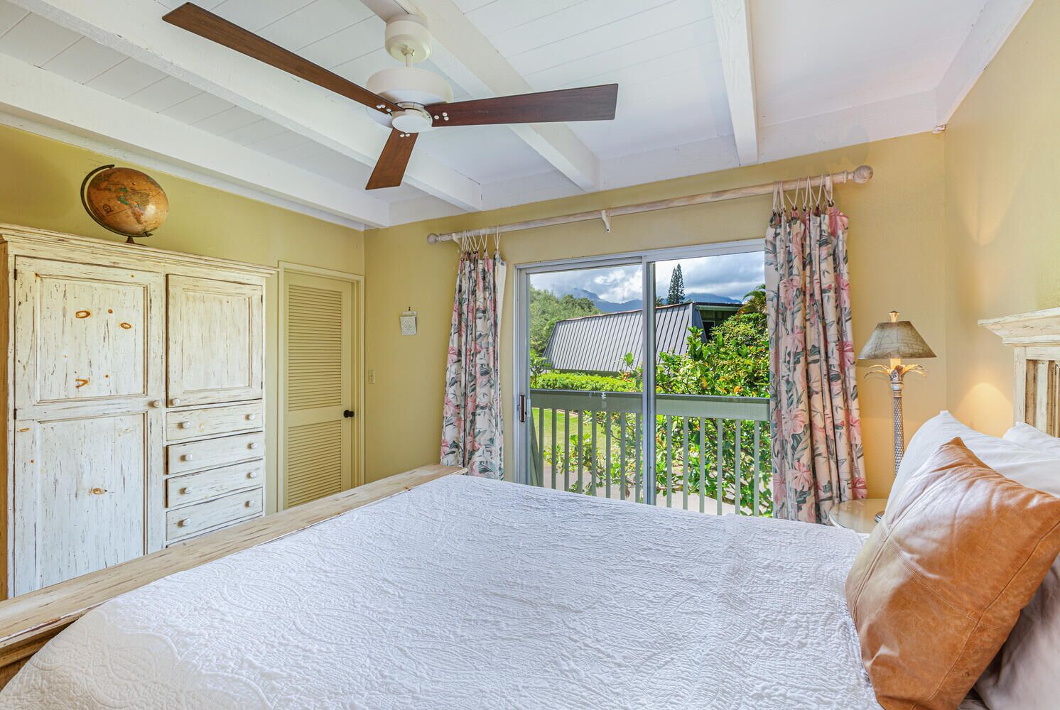 A bedroom with a bed, a ceiling fan, a globe, and a view of a house through a window.