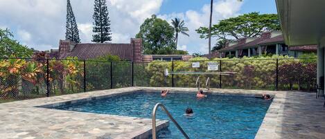 Una piscina con baldosas de piedra, rodeada por una valla, palmeras y un edificio con techo de tejas.