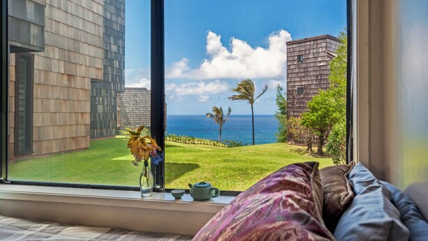 A room with a large window overlooking a grassy area with a view of the ocean and palm trees. - Sealodge F1 (Princeville)