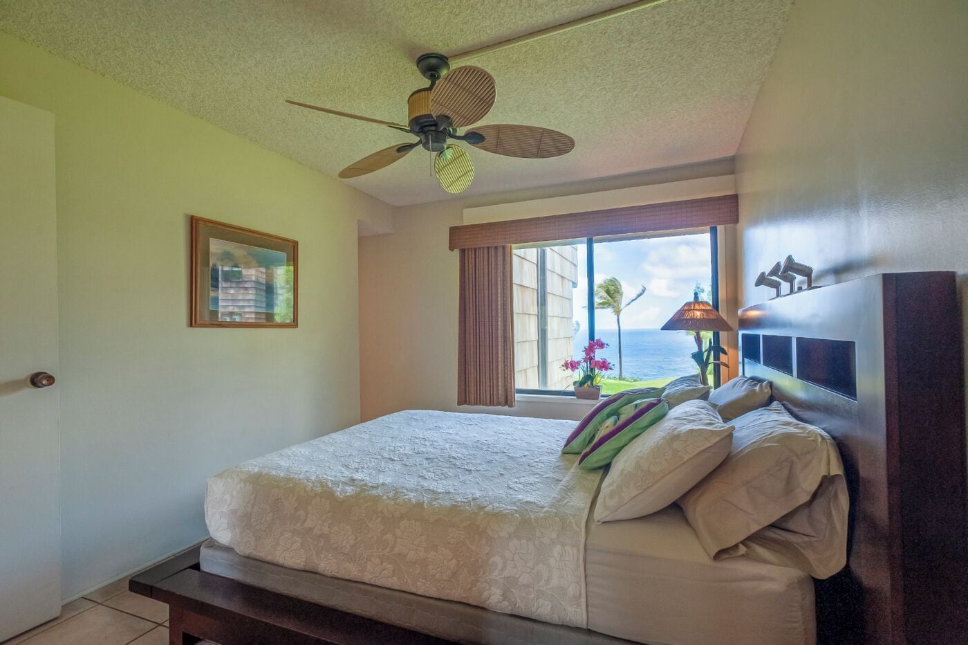 Una camera da letto con un letto, una ventola a soffitto, una finestra vista oceano e un quadro appeso al muro.