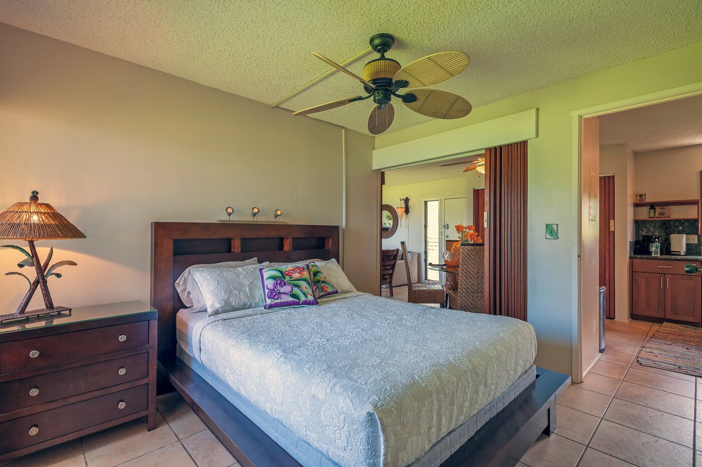 A bedroom with a bed, nightstands, a lamp, a ceiling fan, and a view into the adjacent kitchen.