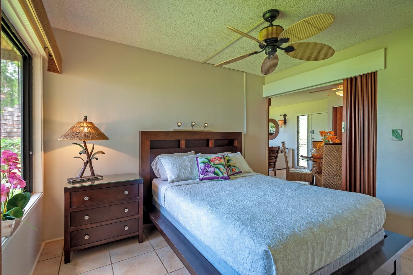 A bedroom with a bed, nightstand, lamp, ceiling fan, and a view of the outside.