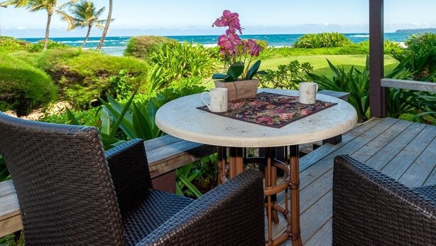 A covered outdoor seating area with wicker chairs, a round table with a floral table runner, and a potted orchid. - Hanalei Colony Resort I-2 (Hanalei)