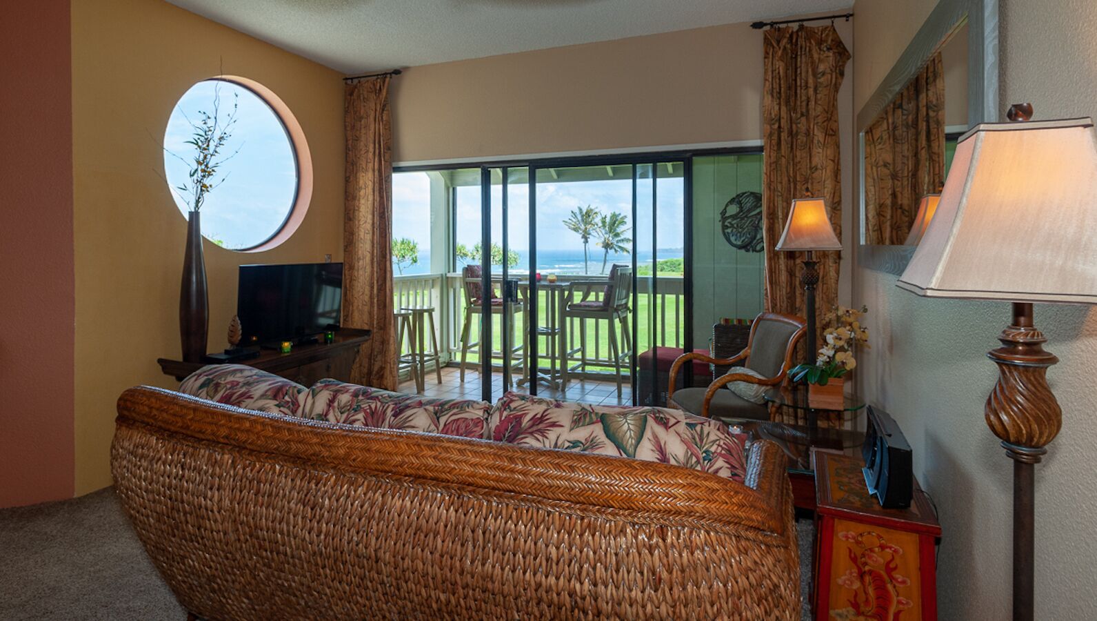 A room with a wicker sofa, a television, a lamp, and a view of a balcony with outdoor furniture.