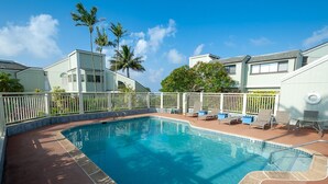 A rectangular swimming pool with a tiled edge, surrounded by a white fence. There are lounge chairs and a table on the poolside. - Kamahana 24 (Princeville)