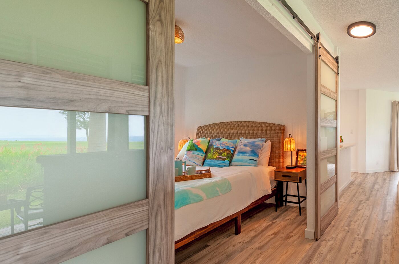 A bedroom with a large bed, a nightstand, a lamp, and a view of the outdoors.