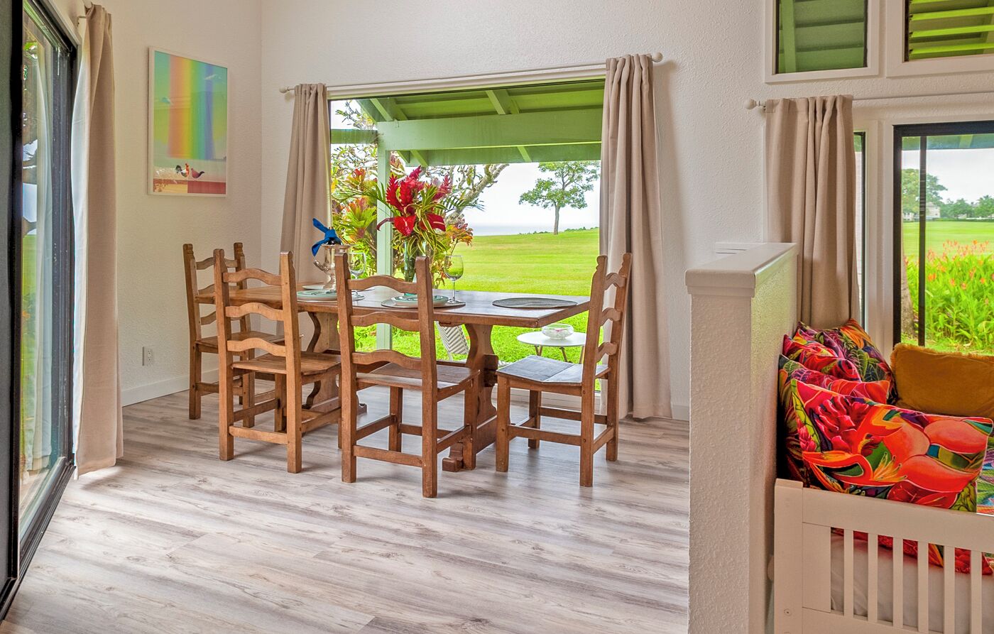 Un comedor con sillas de madera y una mesa, vista a un paisaje verde a través de un ventanal, y un cojín de colores sobre una cuna blanca.