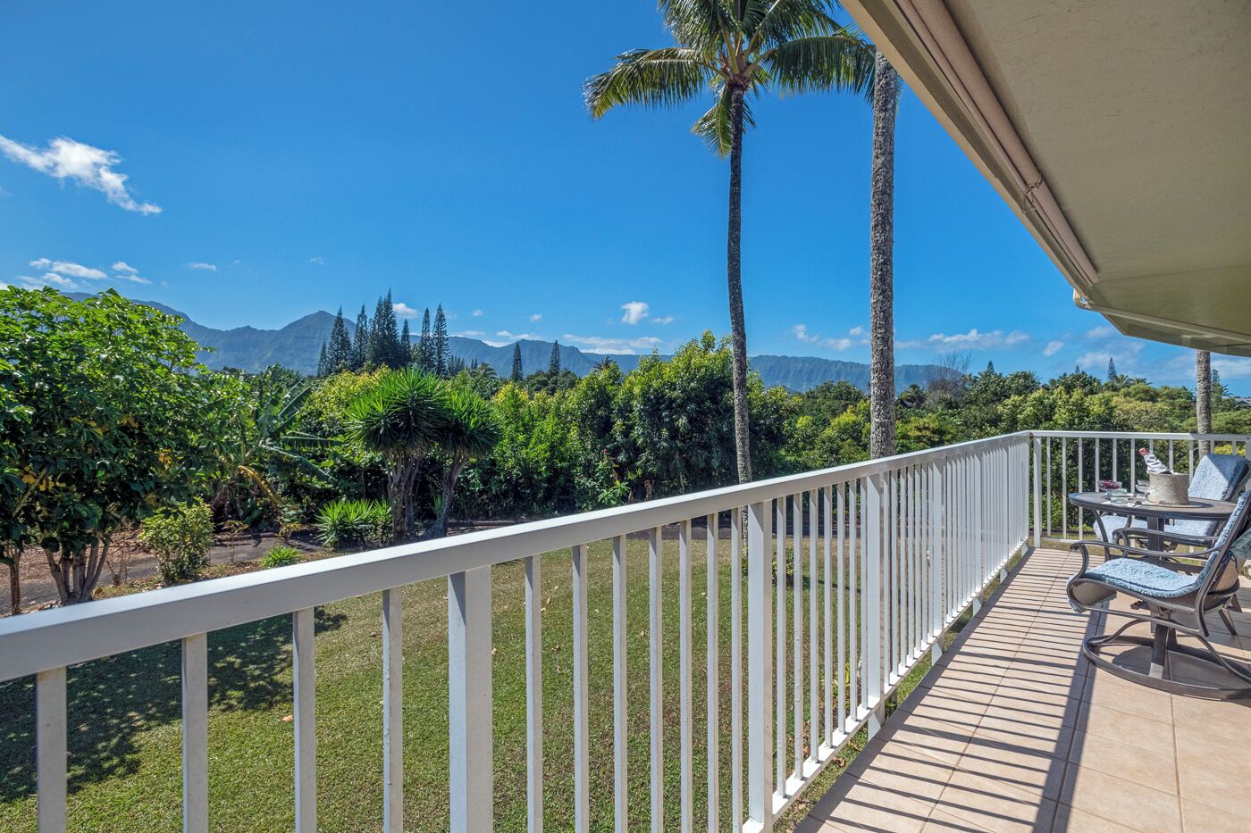 A balcony with a white railing, overlooking a lush garden with palm trees and a mountainous backdrop.