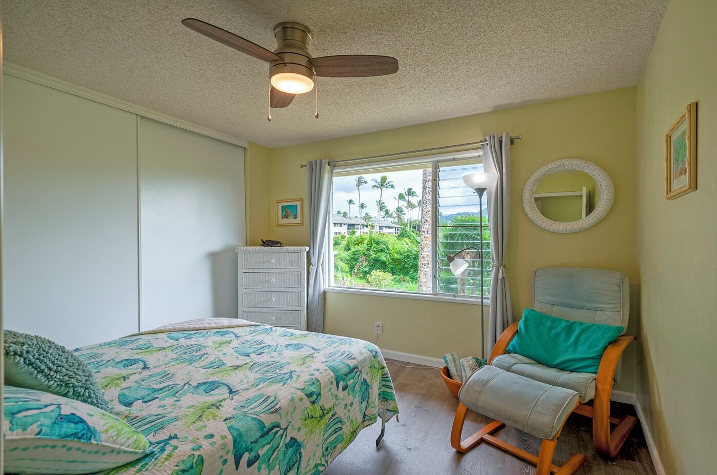 A bedroom with a bed, a chair, a dresser, a window with a view of greenery, and a ceiling fan.