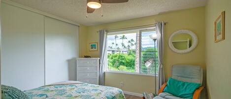 A bedroom with a bed, a chair, a dresser, a window with a view of greenery, and a ceiling fan.