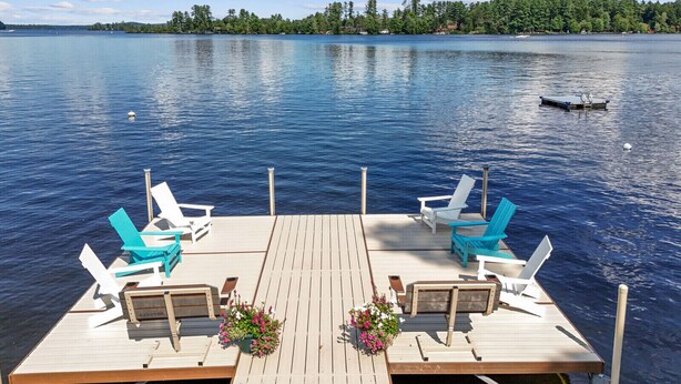 A wooden dock with two blue Adirondack chairs and a table, overlooking a calm lake with a forested shoreline.