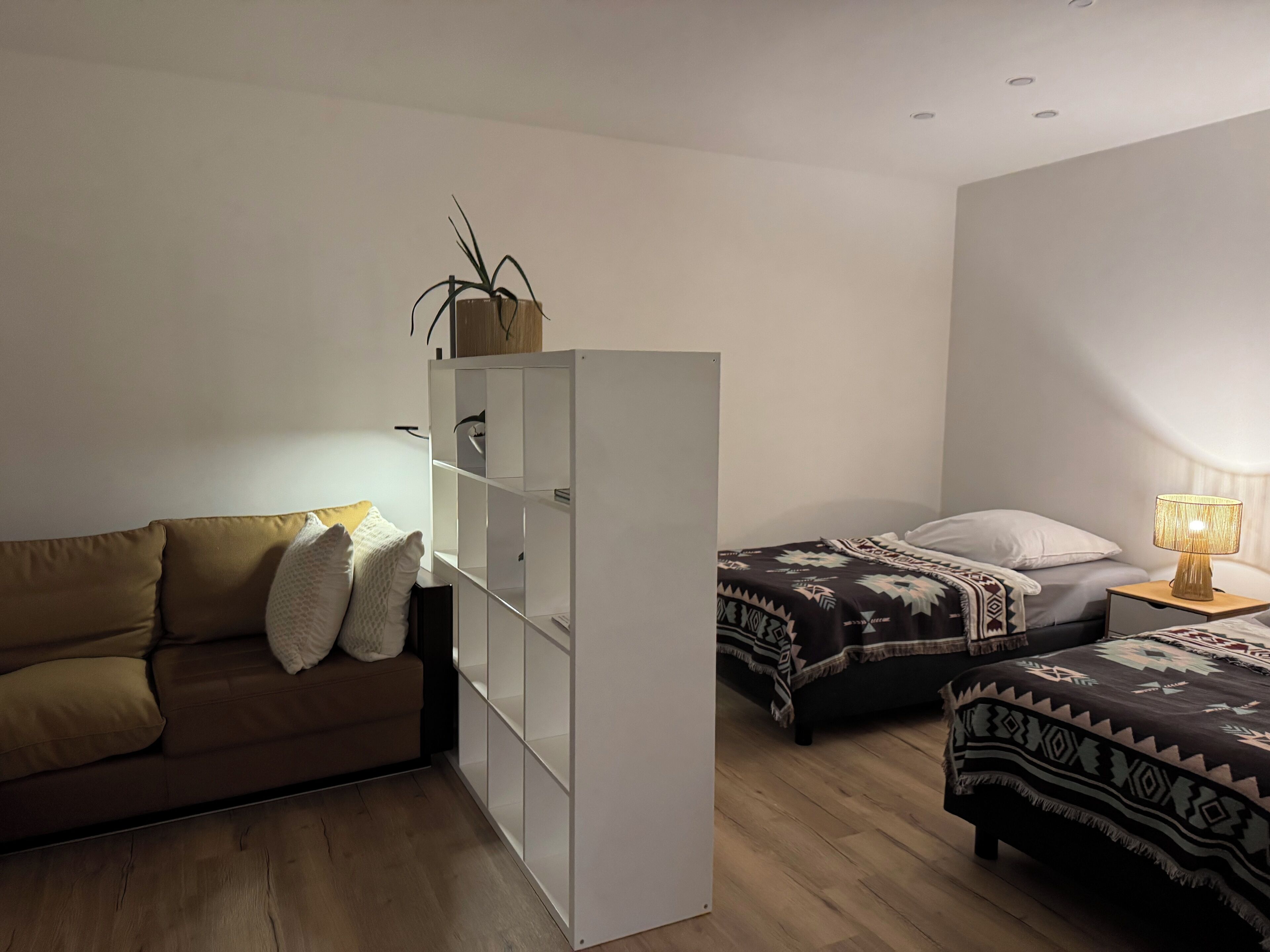 Ein kleiner, gut beleuchteter Raum mit einer Couch, einem Bett mit gemusterten Betttextilien, einem Nachttisch mit Lampe und einem weißen Schrank mit einer Pflanze darauf.