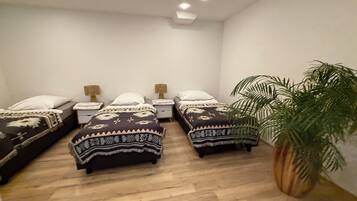 Ein Schlafzimmer mit drei Einzelbetten, jedes mit gemusterten Betttextilien und Nachttischlampen. In der Ecke steht ein großer Blumentopf.