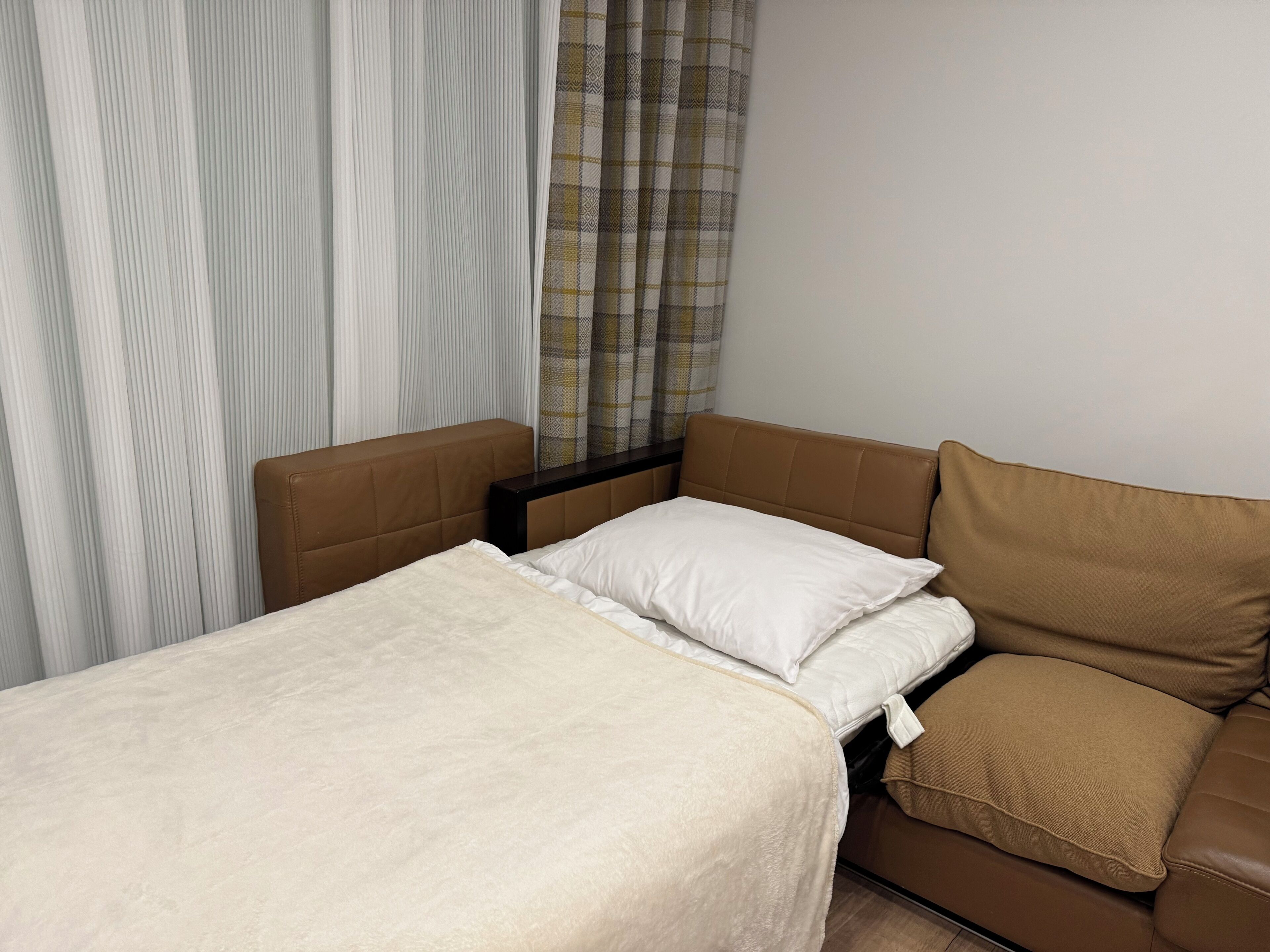 Een kleine, netjes ingerichte kamer met een bed, een bruine bank en een raam met gordijnen.