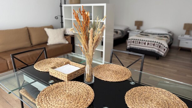 Een moderne woonkamer met een glazen salontafel, geweven onderzetters, een vaas met gedroogde planten, een bruine bank, een witte kast en een bed met een patroon.