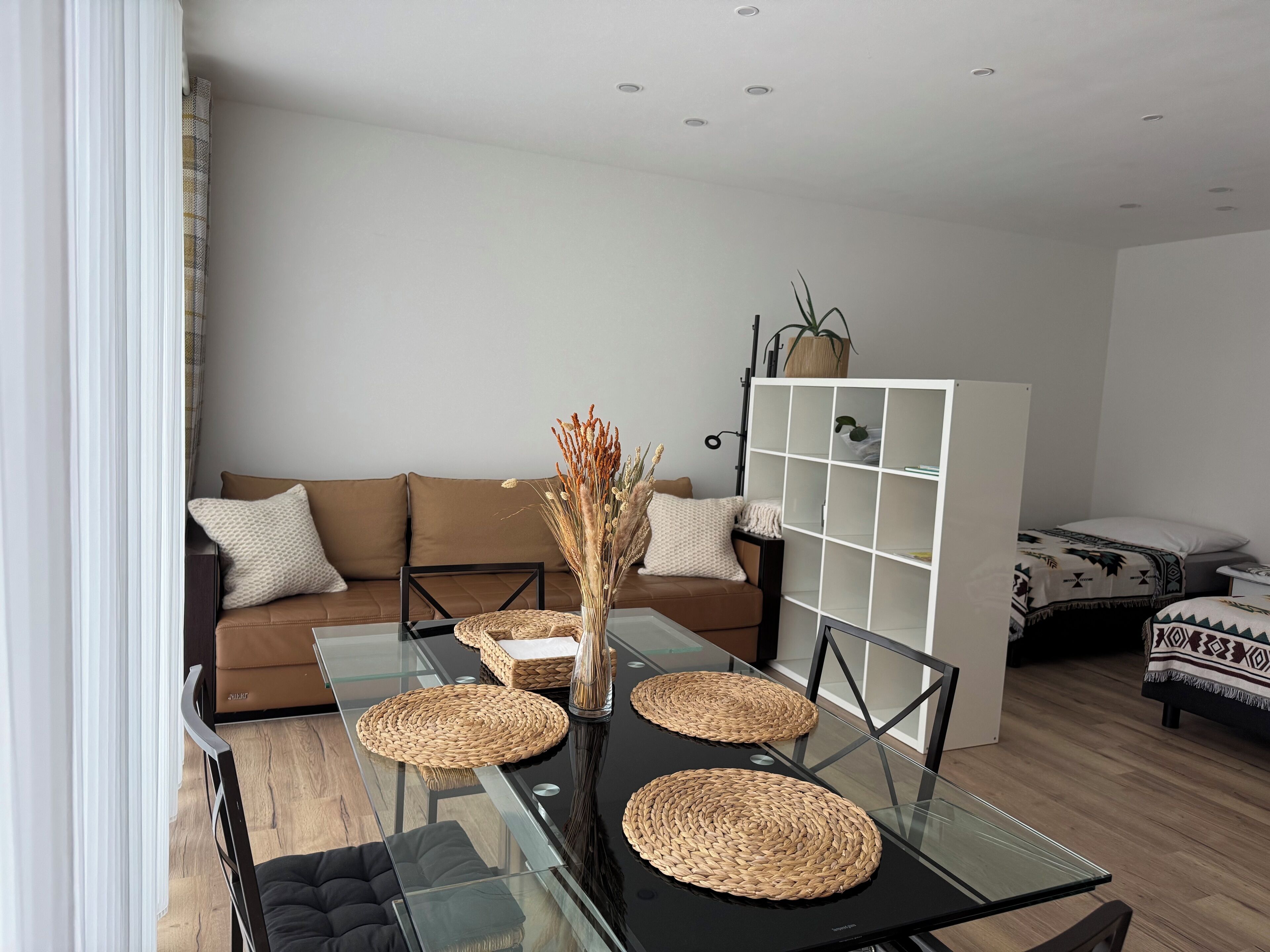 A modern living room with a glass-top dining table, brown sofa, and a white shelving unit.