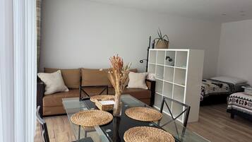 A modern living room with a glass-top dining table, brown sofa, and a white shelving unit.