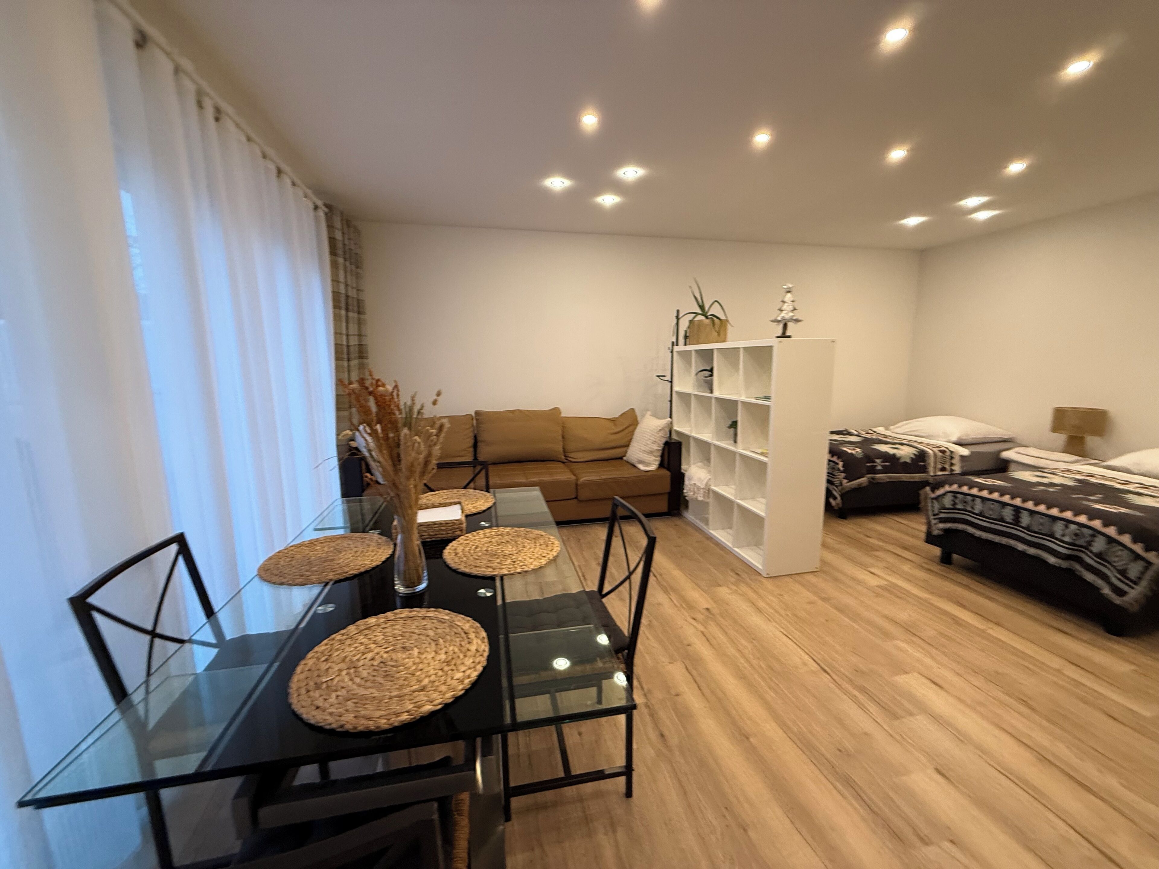 A modern living room with a glass dining table, a sofa, a bookshelf, and a bed.