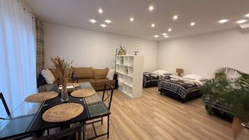 A modern living room with a glass dining table, a sofa, a bookshelf, and two beds.