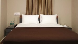 Habitación de hotel con una cama grande, dos almohadas blancas, un cabecero marrón, una mesita de noche con un teléfono y botellas de agua, y un equipo de aire acondicionado en la pared.